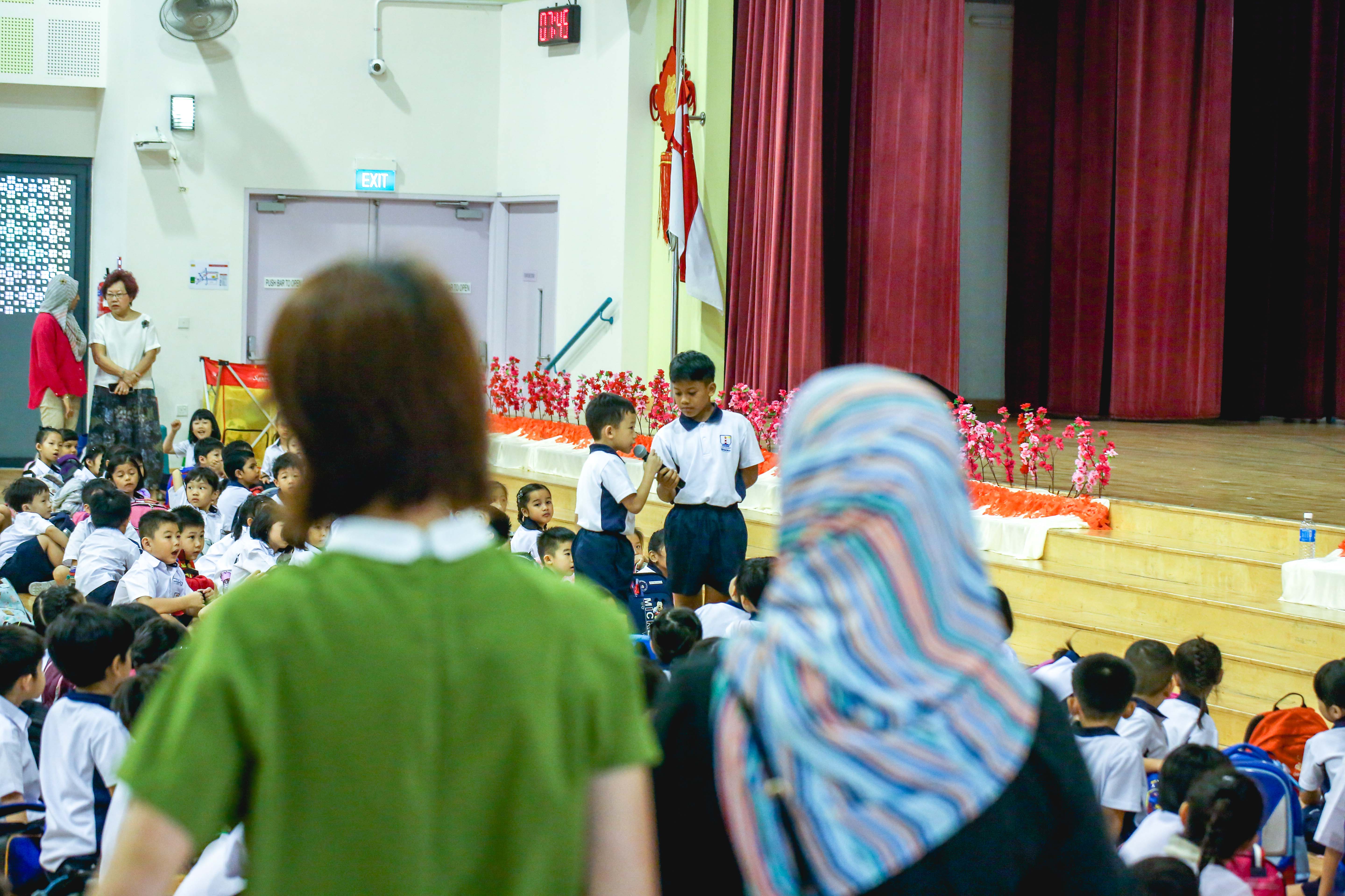 Two young students speak into a microphone on a school hall stage while uniformed children sit on the floor watching.